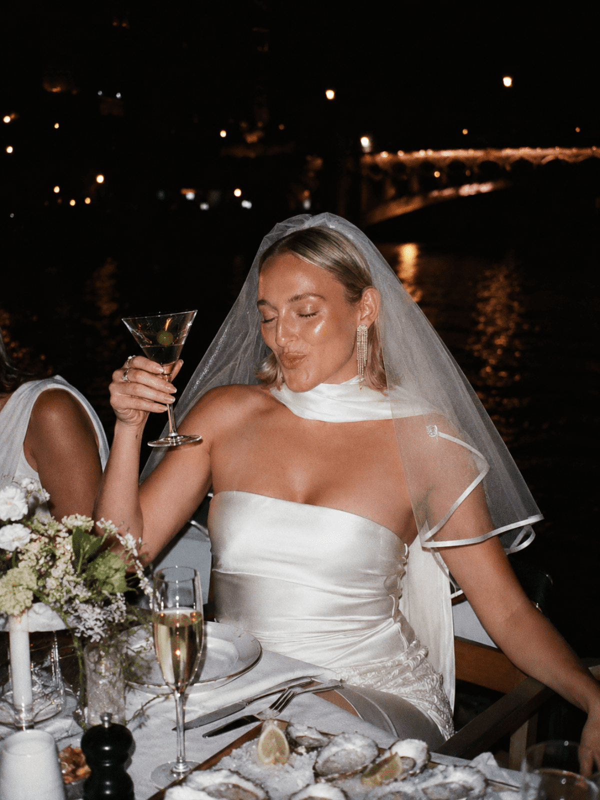 Newcastle hen do activities inspiration image of a bride-to-be in a veil holding a martini at a candlelit dinner, with flowers, champagne and city lights in the background.
