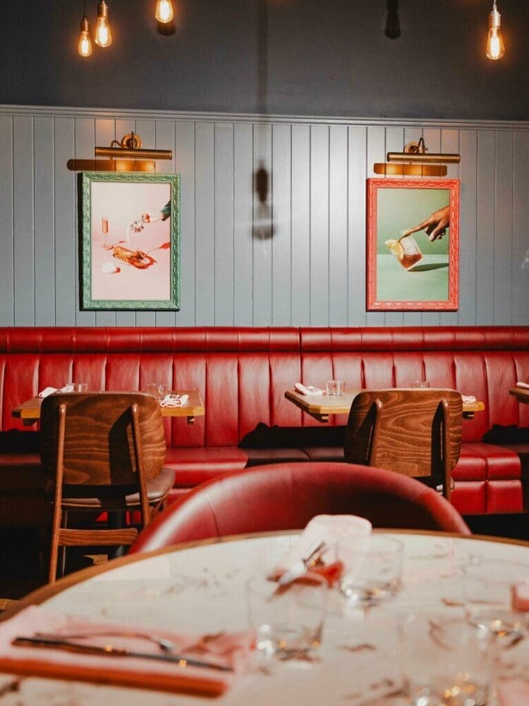 The stylish and vibey interior of Burger and Lobster restaurant, featuring long, plush red leather booth seating against a paneled light blue-grey wall. Two illuminated art prints are hung above the booths: the left depicts a lobster and cocktails on a pink background, while the right shows a hand holding a refreshing drink. The warm glow from exposed Edison bulbs adds to the inviting atmosphere, making it an ideal spot for a fun hen weekend dinner in Brighton.