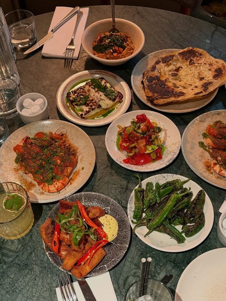 A vibrant spread of delicious sharing plates and a cocktail on a dark green table at Burnt Orange restaurant in Brighton. This selection of tapas-style dishes creates a perfect vibey dinner experience for a hen weekend, featuring prawns, flatbread, grilled halloumi, and more.