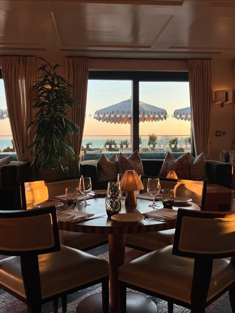 The elegant dining room of Cecconi's in Brighton, bathed in a warm golden hour glow. A beautifully set table for guests is in the foreground, with plush seating and a small, intimate lamp. Through large windows, the sparkling sea and the charming white scalloped umbrellas of an outdoor terrace are visible, creating a luxurious and truly vibey ambiance ideal for a memorable hen weekend dinner.