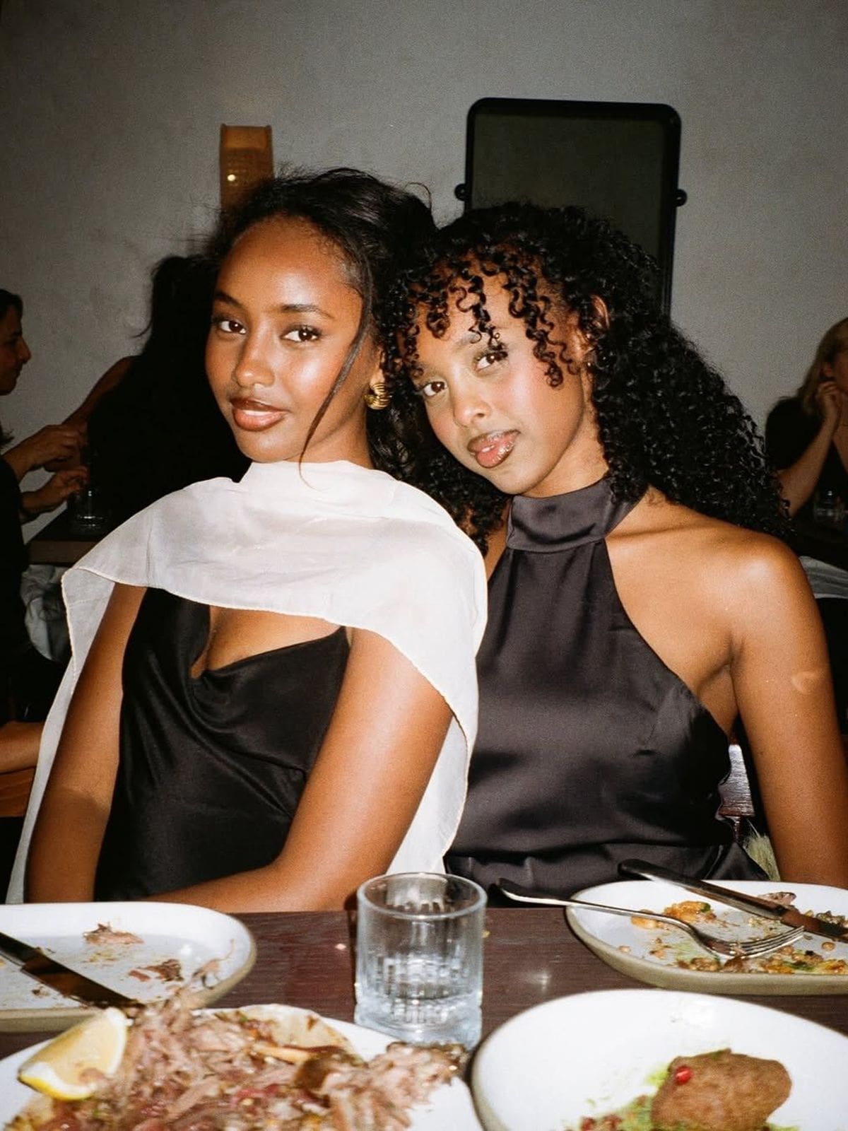 Two women sitting close together at a restaurant table, dressed up for a night out in satin tops. Plates of finished food and a glass of water sit in front of them. The photo has a warm, film-camera feel with soft lighting and other diners visible in the background.