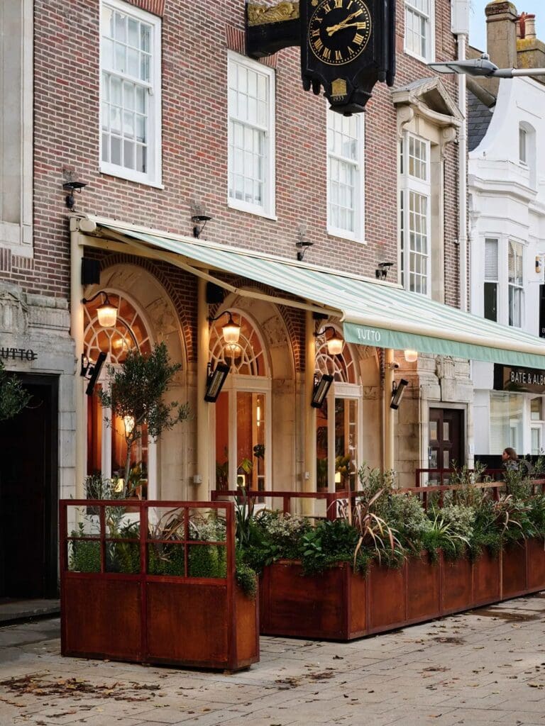 The charming brick exterior of Tutto restaurant in Brighton, featuring an inviting outdoor terrace with planters and a green-striped awning. Large arched windows reveal the warm glow of the interior, topped with a vintage-style clock, making it an elegant and vibey choice for a hen weekend dinner.