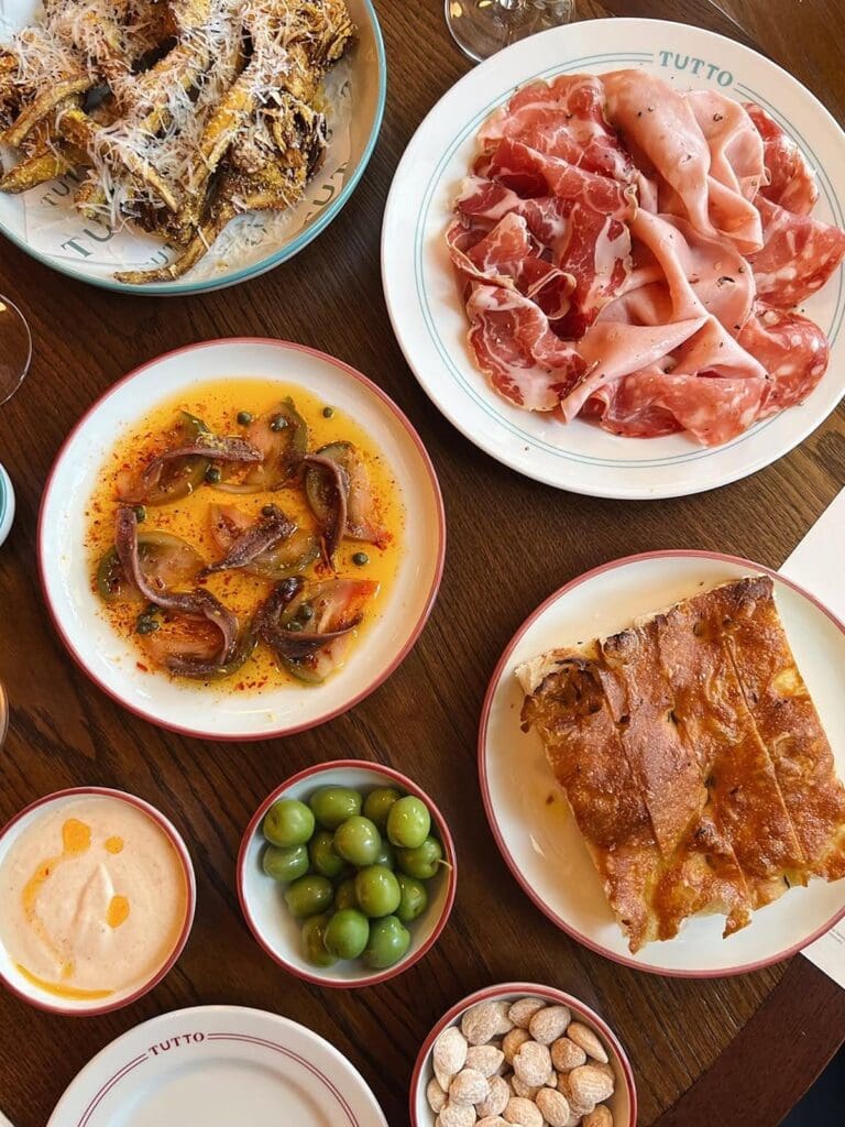 An overhead shot of a feast of Italian sharing plates on a wooden table at Tutto restaurant. Dishes include platters of cured meats, anchovy and caper salad, focaccia, fried zucchini with parmesan, olives, and various dips, creating a delicious and vibey spread for a hen weekend in Brighton.