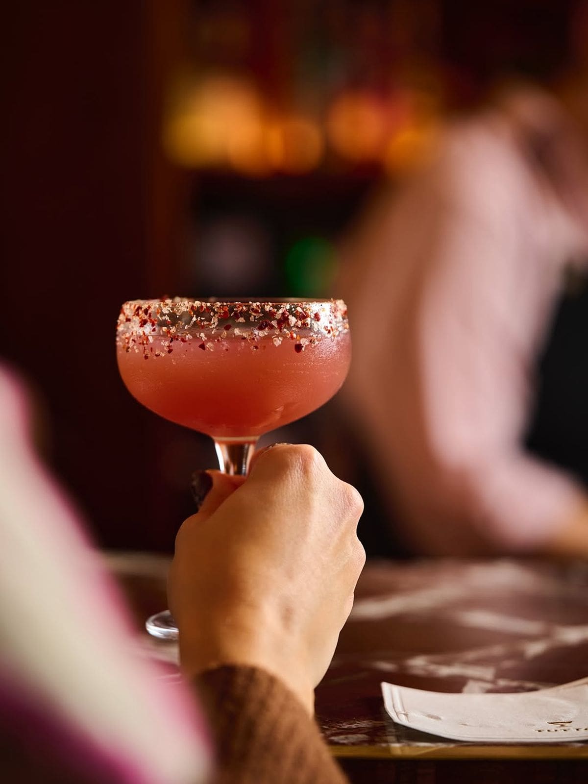 A hand holding a vibrant pink cocktail with a chili-salt rim at Tutto restaurant, with a blurred bar scene in the background. This stylish drink sets a perfectly vibey tone for a hen weekend dinner in Brighton.