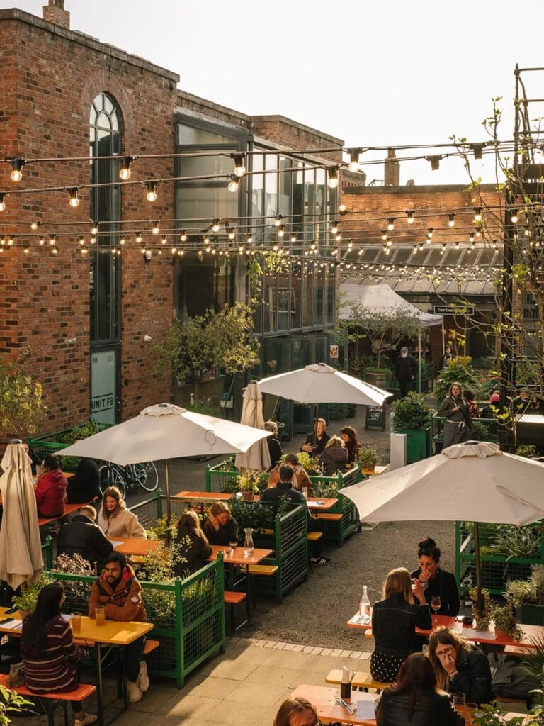 The sunny courtyard at Buyers Club in Liverpool, with groups of people eating and drinking at orange-topped tables surrounded by green metal planters, white parasols, festoon lights strung overhead, and a converted red brick warehouse with large steel-framed windows in the background.
