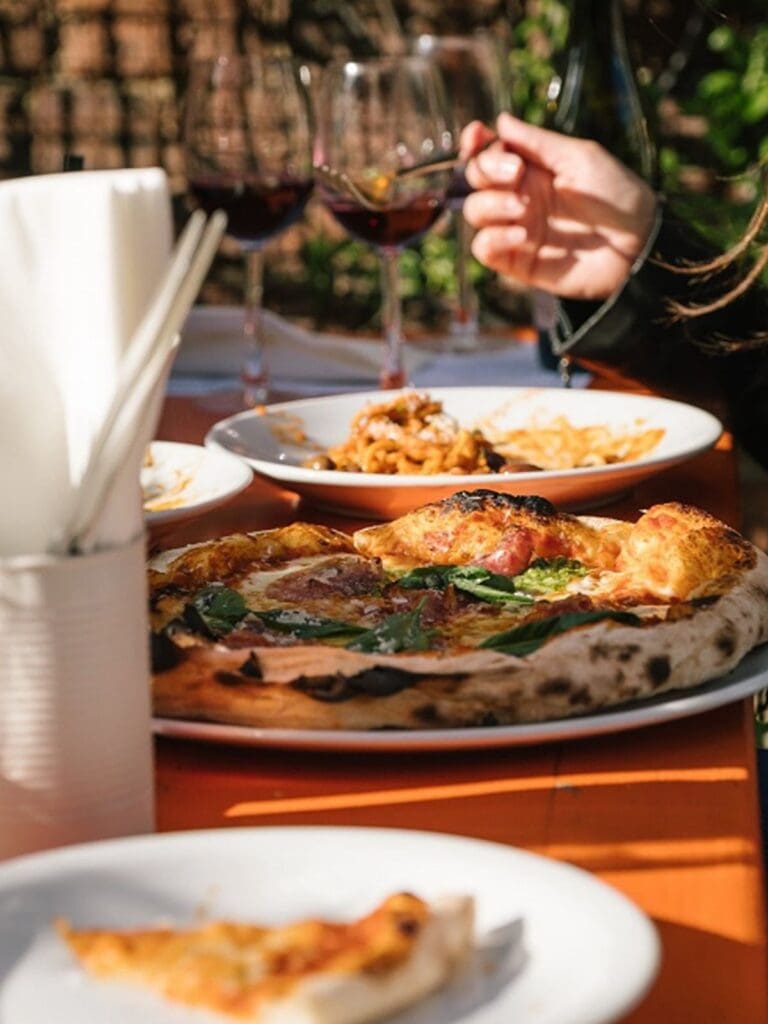 A close-up of a wood-fired pizza topped with spinach and cured meat alongside a bowl of pasta and two glasses of red wine on an orange table in the sunshine at Buyers Club in Liverpool.