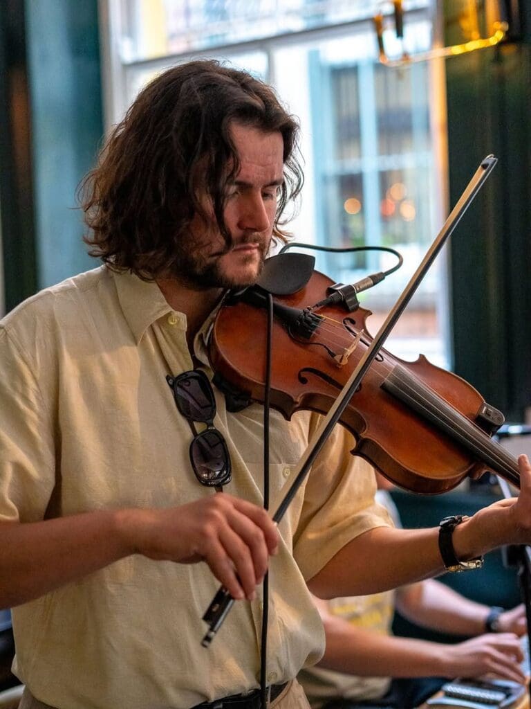 A musician playing the violin during a live music session at Castle Street Townhouse in Liverpool, wearing a linen shirt with sunglasses hanging from the collar, with warm natural light coming through the window behind him.