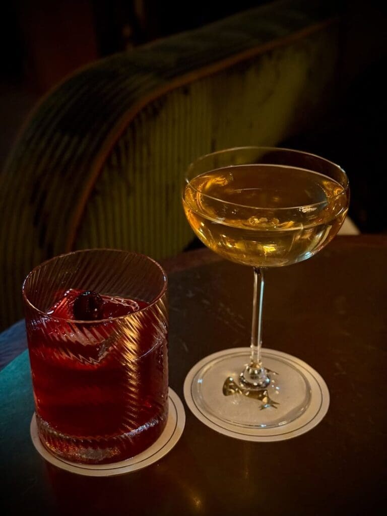 Two cocktails on coasters at Hawksmoor in Liverpool — a deep red negroni with a cherry in a textured glass tumbler and a golden cocktail in a coupe glass, set on a dark wooden table with moody, low lighting.