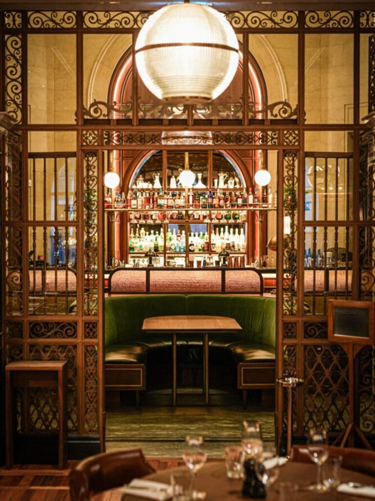 A green velvet booth framed by ornate wrought iron screens and a large globe pendant light at Hawksmoor in Liverpool, with the fully stocked bar visible through the archway behind and a set dining table in the foreground.