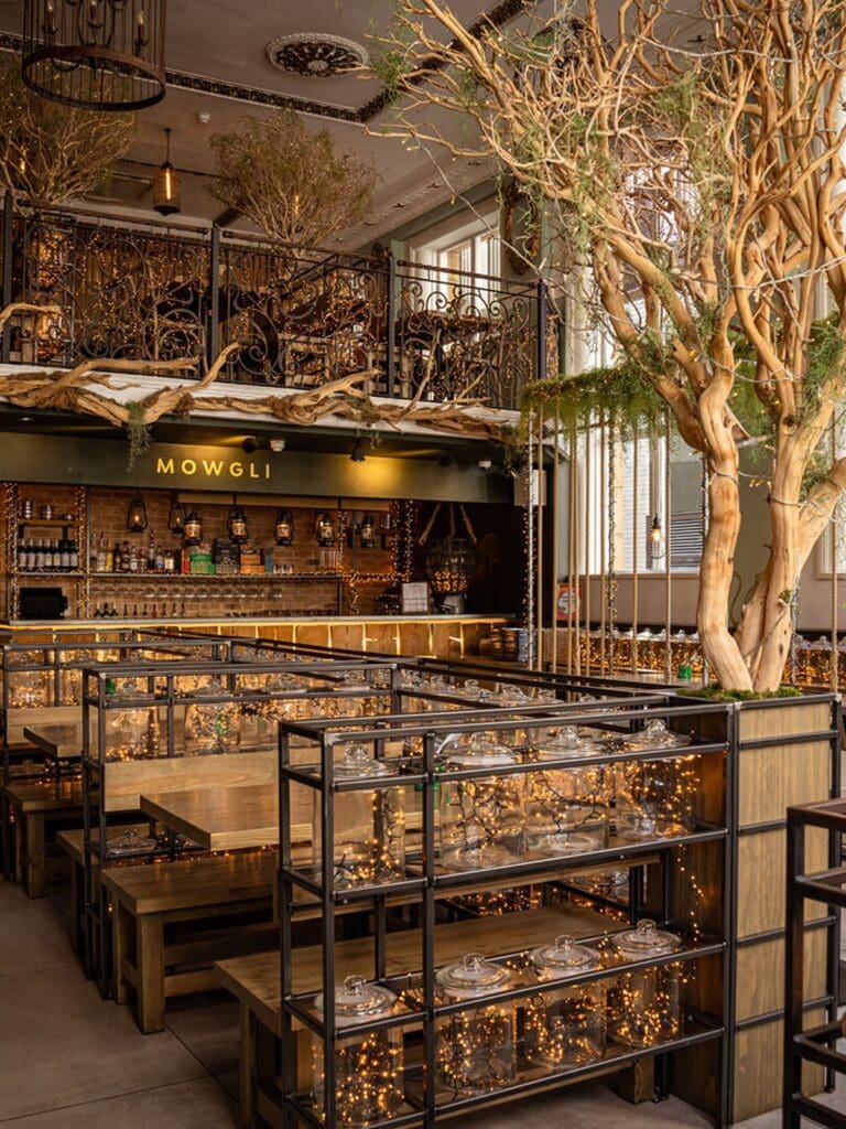 The interior of Mowgli in Liverpool, with wooden booth seating divided by black metal shelving units filled with glass cloches and fairy lights, a large statement tree stretching up through the double-height space, ornate wrought iron balcony railings on the mezzanine level, and the warmly lit bar with lanterns and exposed brick in the background.