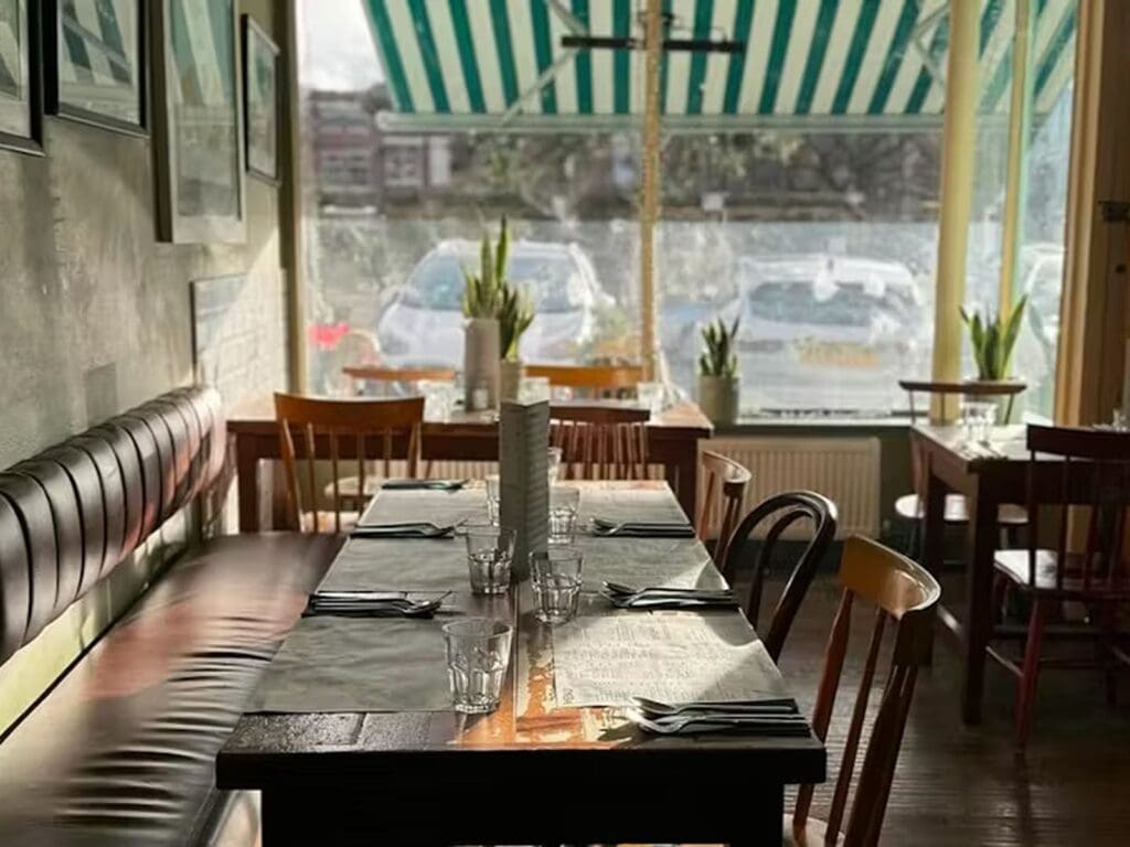 A sunlit table set for a group at The Quarter in Liverpool, with leather banquette seating, dark wooden chairs, water glasses and cutlery on slate placemats, potted plants on the windowsill and a green and white striped awning visible through the large front window."