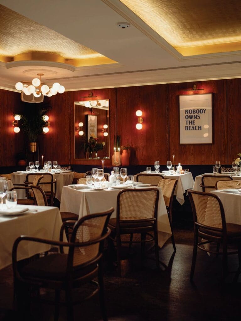 Candlelit dining room at Bagatelle London with white tablecloths, warm wood-panelled walls and intimate lighting — a glamorous dinner spot for a London hen weekend.