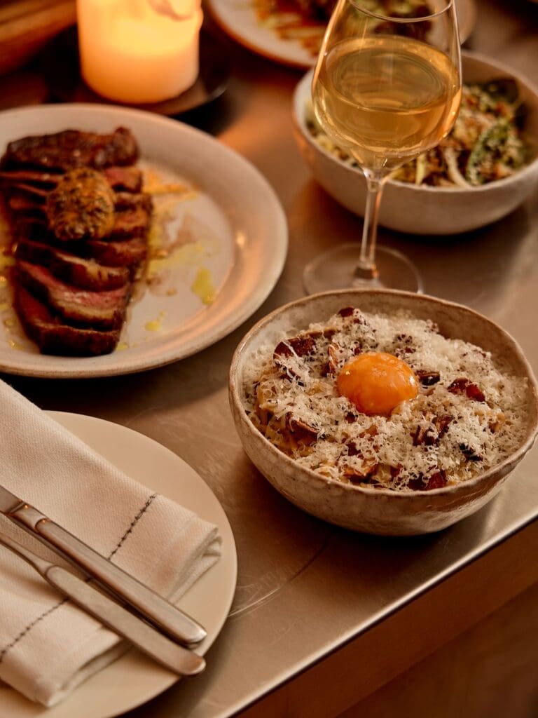 Plates of pasta topped with egg yolk and parmesan, sliced steak and a glass of white wine at Bambi in London.