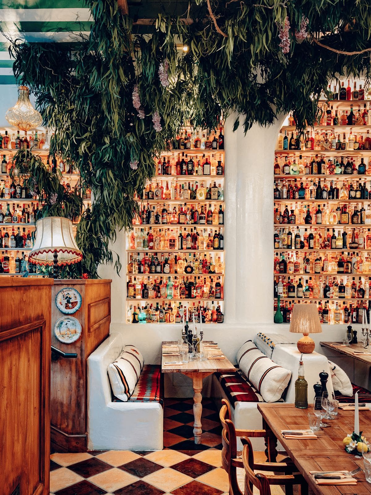 Circolo Popolare London dining room with cascading greenery overhead, floor-to-ceiling shelves of colourful spirit bottles and cosy booth seating set for a lively group dinner.