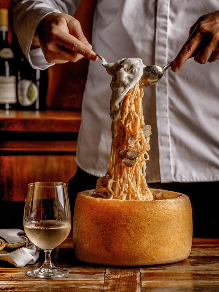 Truffle spaghetti tossed in a giant wheel of cheese at Gloria in London, served tableside with a glass of white wine in a buzzy Italian restaurant setting.