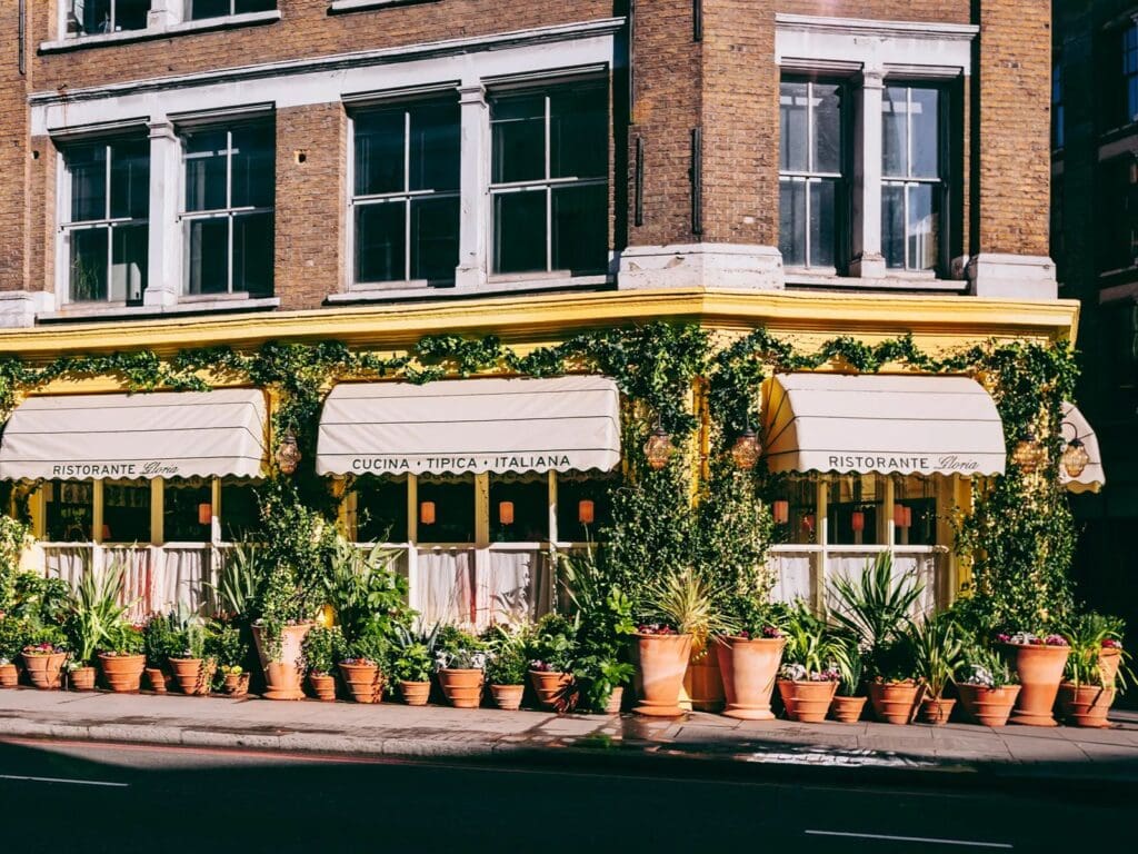 Exterior of Gloria restaurant in London with yellow façade, striped awnings and rows of terracotta plant pots lining the entrance.