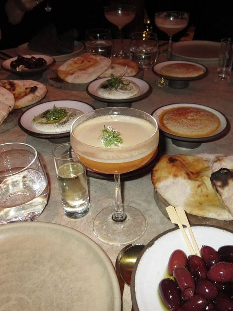 Sharing plates and cocktails at Fenix Manchester including flatbreads, dips, olives and a coupe cocktail, photographed on a marble table during a lively group dinner.
