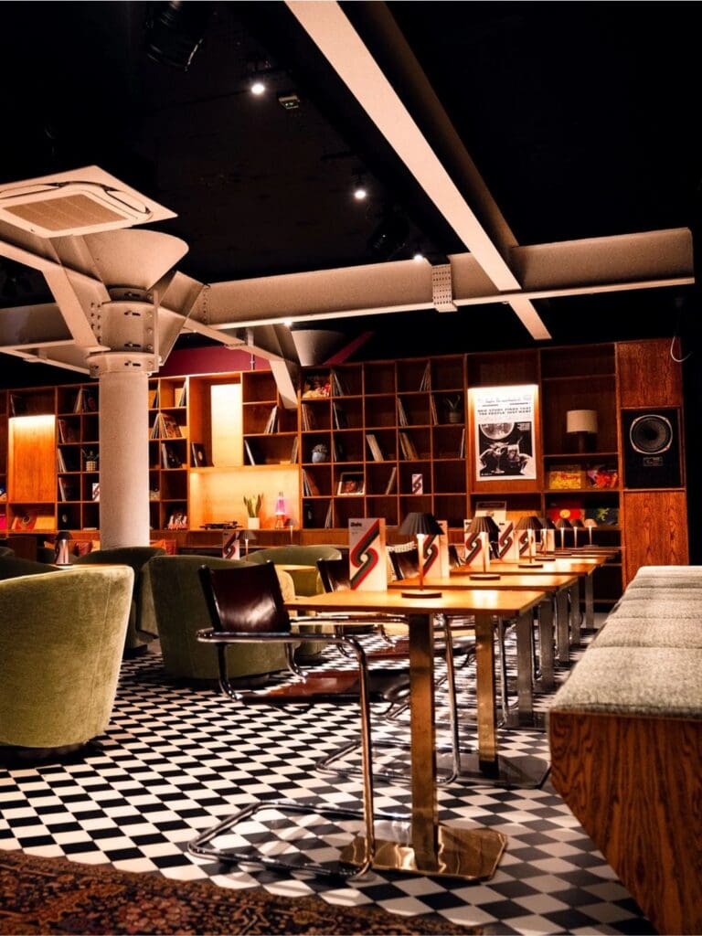 Dining room at Tartuffe at Side Street Manchester featuring record-lined shelving, checkerboard flooring, soft table lamps and low-lit seating for a stylish evening dinner.