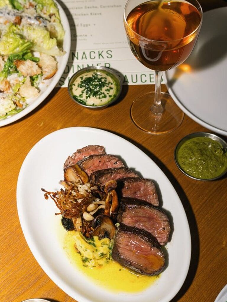 Sliced medium-rare steak with mushrooms and butter sauce served at Tartuffe at Side Street in Manchester, photographed with side dishes and a glass of rosé wine during a vibey dinner.