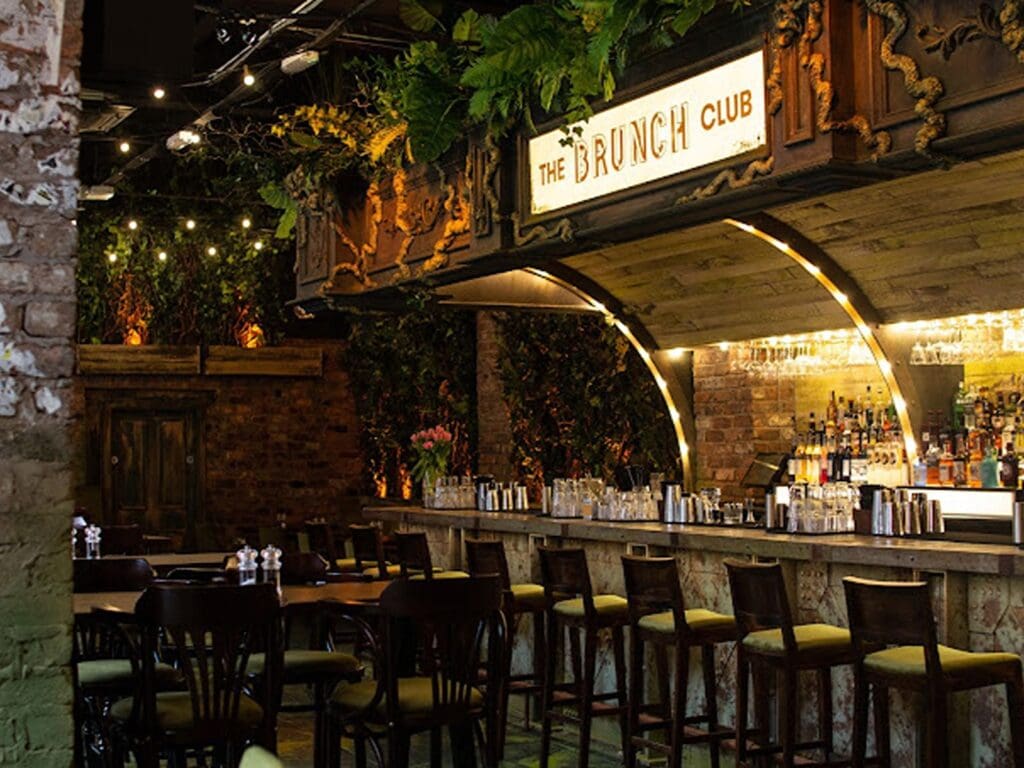 Moody bar interior at The Brunch & Cocktail Club Liverpool, with “The Brunch Club” sign, foliage-draped ceiling and high stools — a chic hen weekend brunch spot.