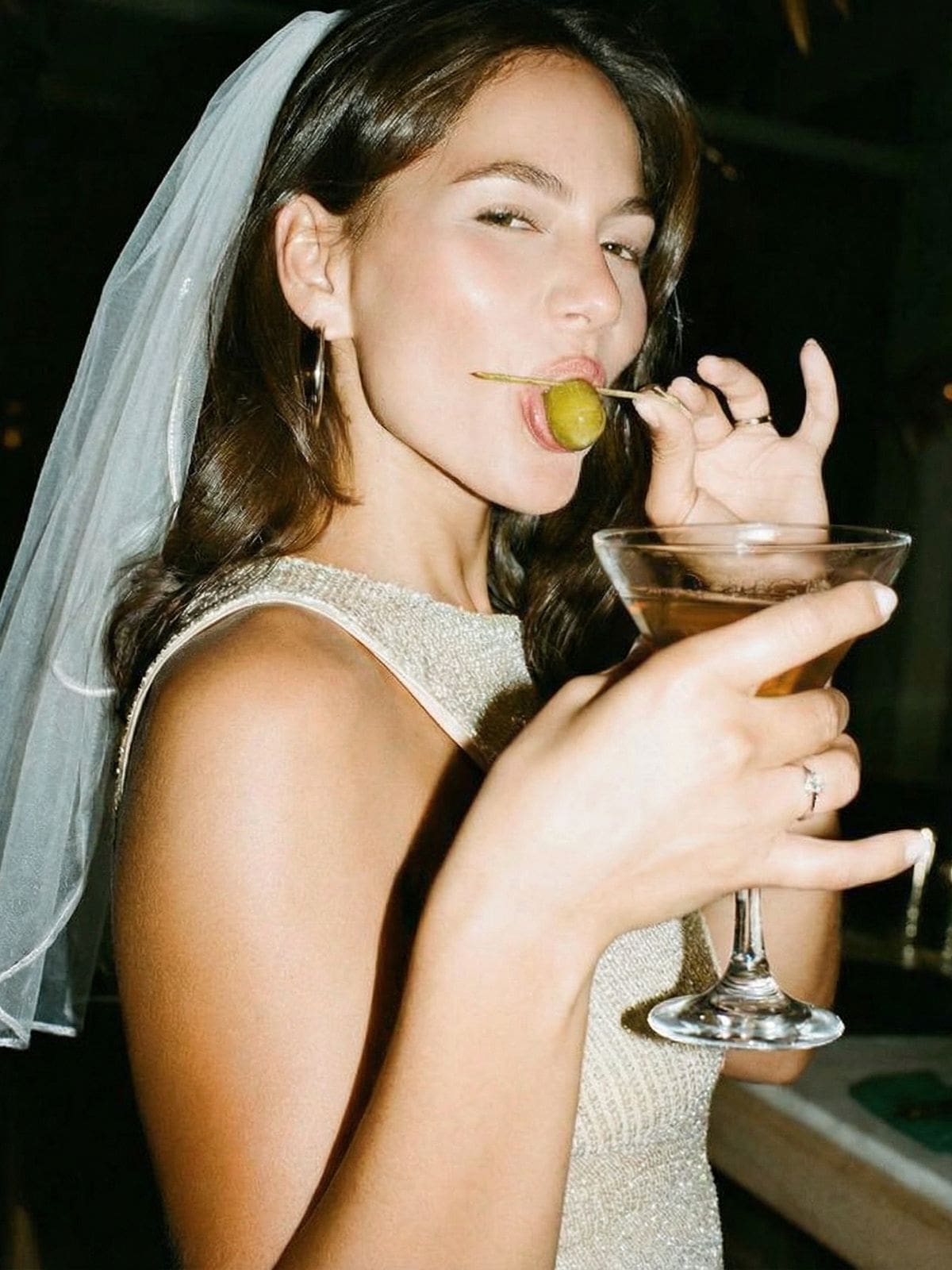 A bride wearing a veil holds a martini and playfully bites an olive, capturing a glamorous hen weekend moment during a boozy brunch.