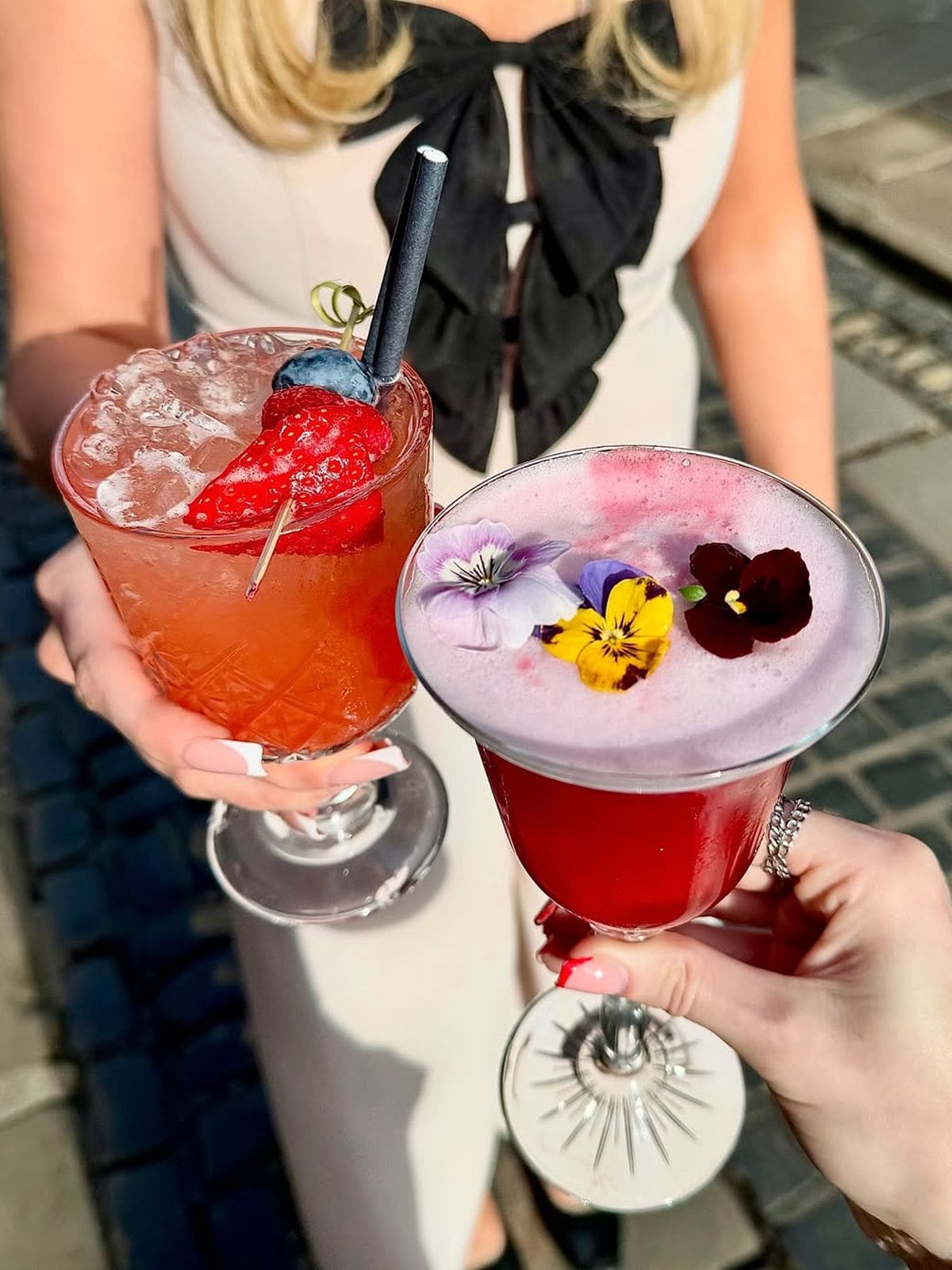 Two colourful cocktails held up in the sunshine outside Lucia York, garnished with berries and edible flowers, perfect girls’ weekend moment.