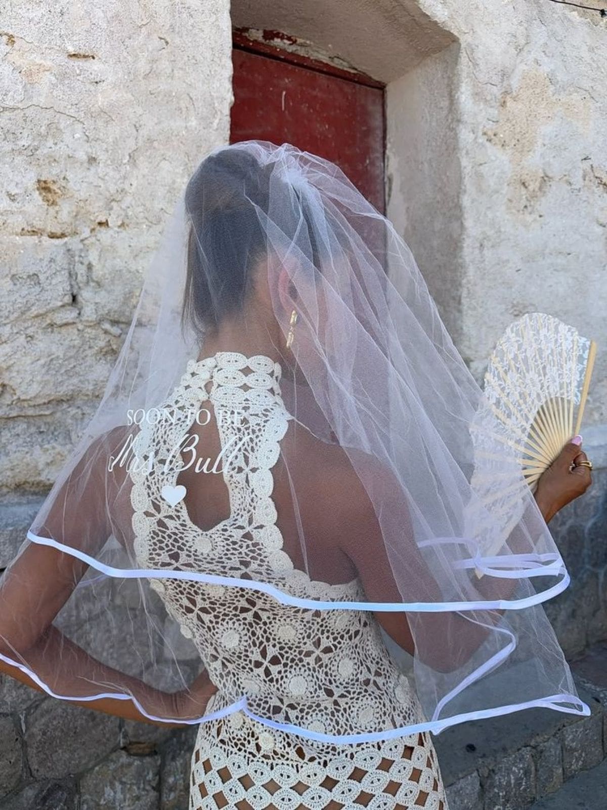 Lisbon hen activities inspiration image of a bride-to-be in a white crochet dress and veil, holding a lace fan against a rustic stone wall.