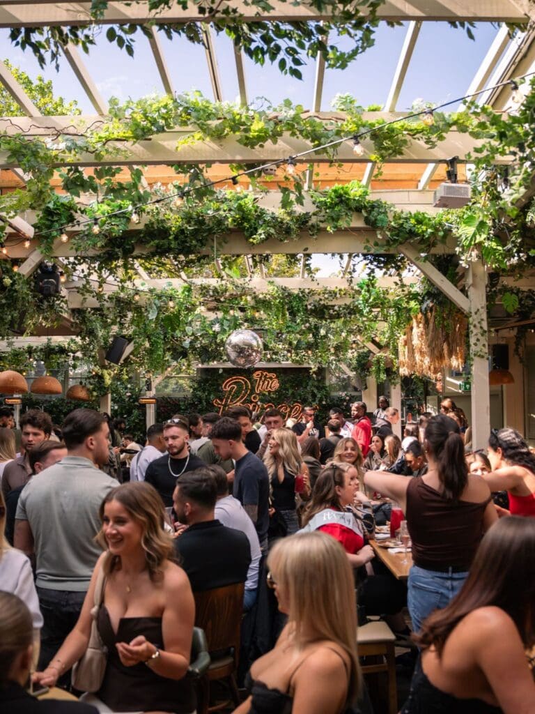 A packed bottomless brunch at The Prince, London, in the covered outdoor-indoor courtyard, with long tables, hanging greenery and a full-on weekend buzz.