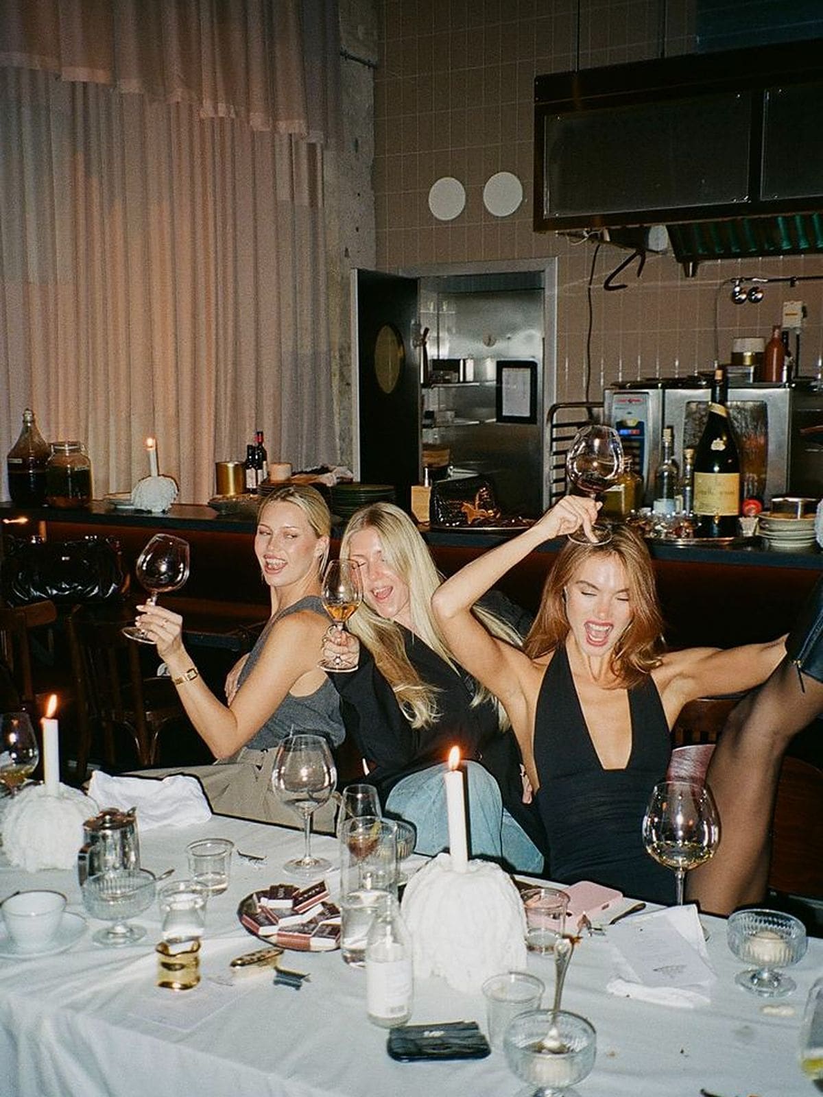 Three women laughing and raising their wine glasses at the end of a big dinner, sitting at a white tablecloth-covered table scattered with candles, empty glasses and plates. The photo has a warm, flash-lit film camera feel with a restaurant kitchen visible in the background.