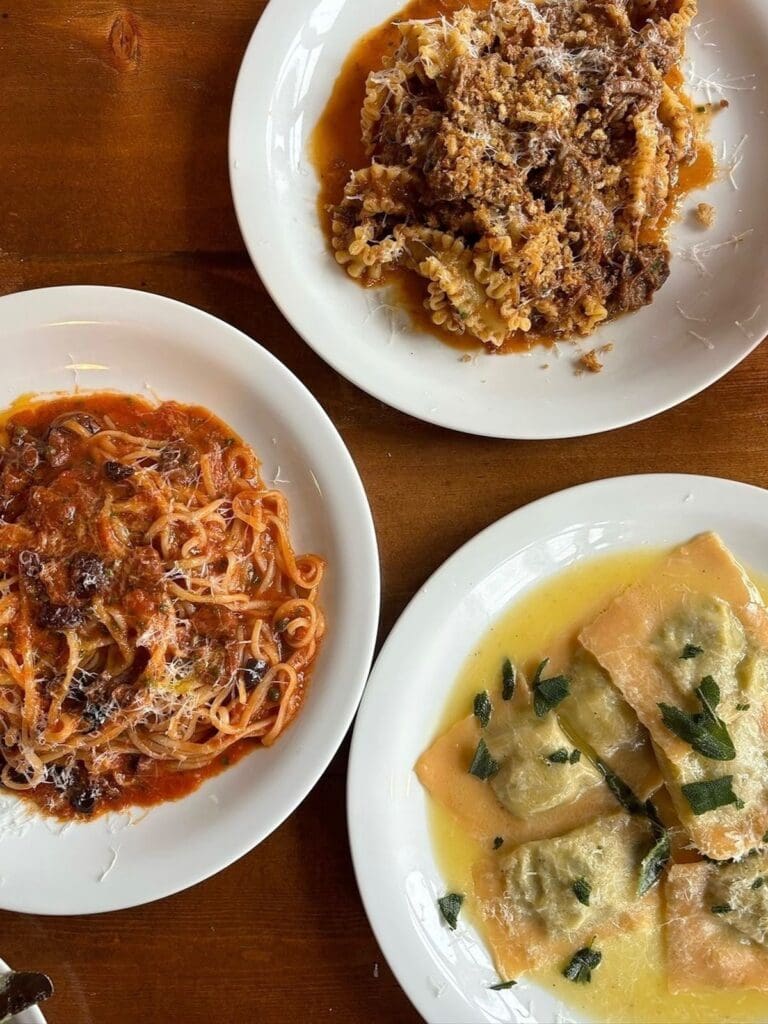 An overhead shot of three pasta dishes on white plates at Solina in Bath — a spaghetti in rich tomato sauce, a fusilli with a slow-cooked meat ragù topped with breadcrumbs and parmesan, and ravioli in a butter and sage sauce, all served on a dark wooden table.