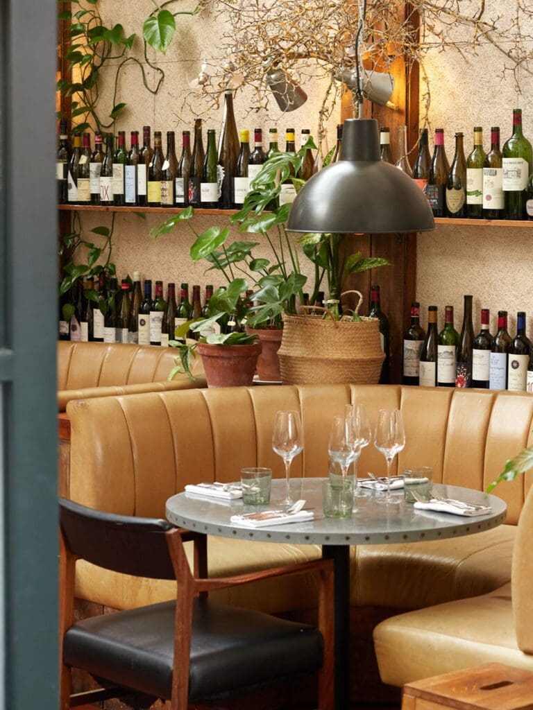 A cosy corner booth at Walcot House in Bath with tan leather banquette seating, a round marble table set with wine glasses and cutlery, shelves of wine bottles lining the wall and potted plants dotted throughout