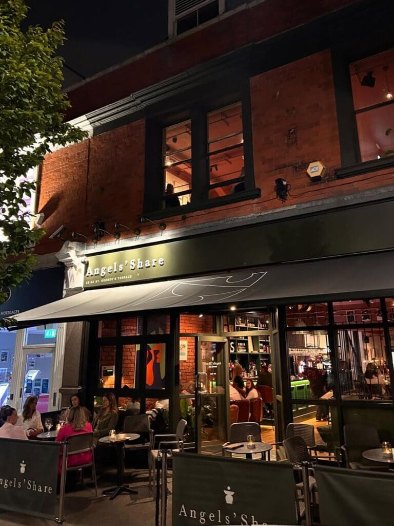 The exterior of Angels' Share in Jesmond, Newcastle at night, with a dark green branded awning, warm light glowing through the large glass-fronted windows, groups of women sitting at candlelit tables on the terrace, and the busy bar visible inside