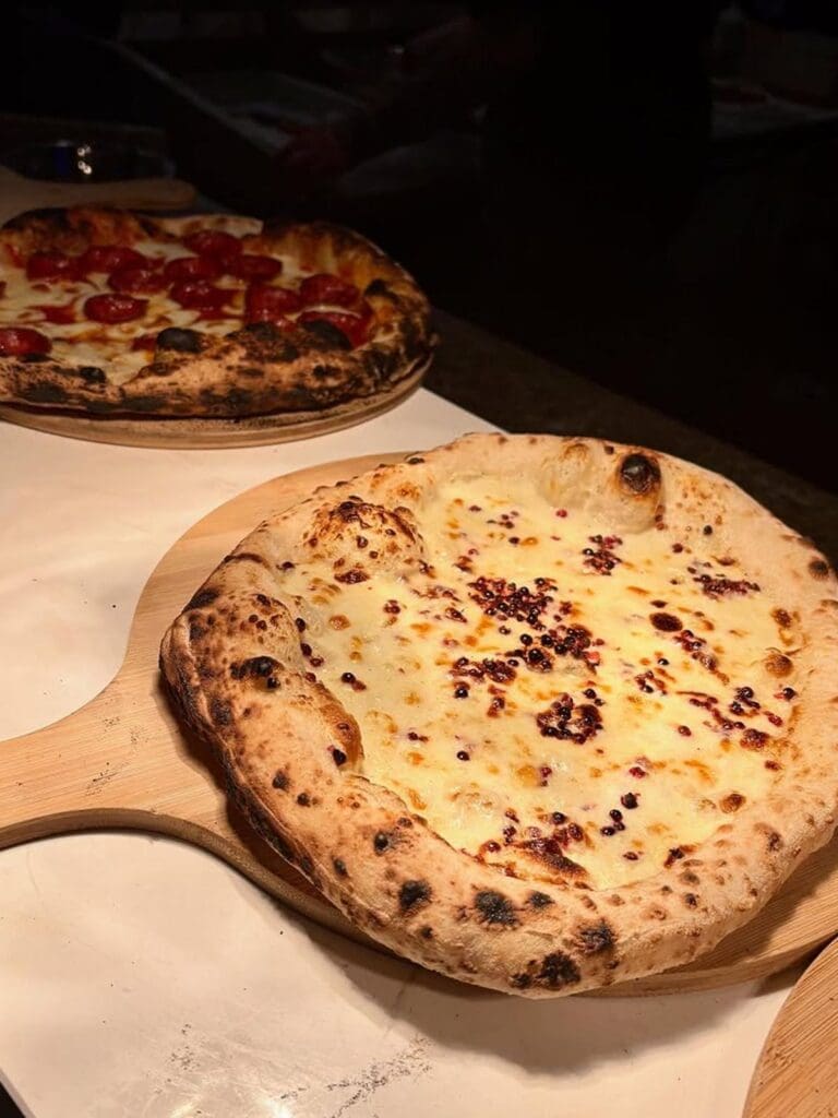 Two freshly baked Neapolitan pizzas on wooden paddles at the pass at Sanco in Newcastle — one topped with melted cheese and pink peppercorns, the other with cherry tomatoes and mozzarella, both with charred, puffy crusts.