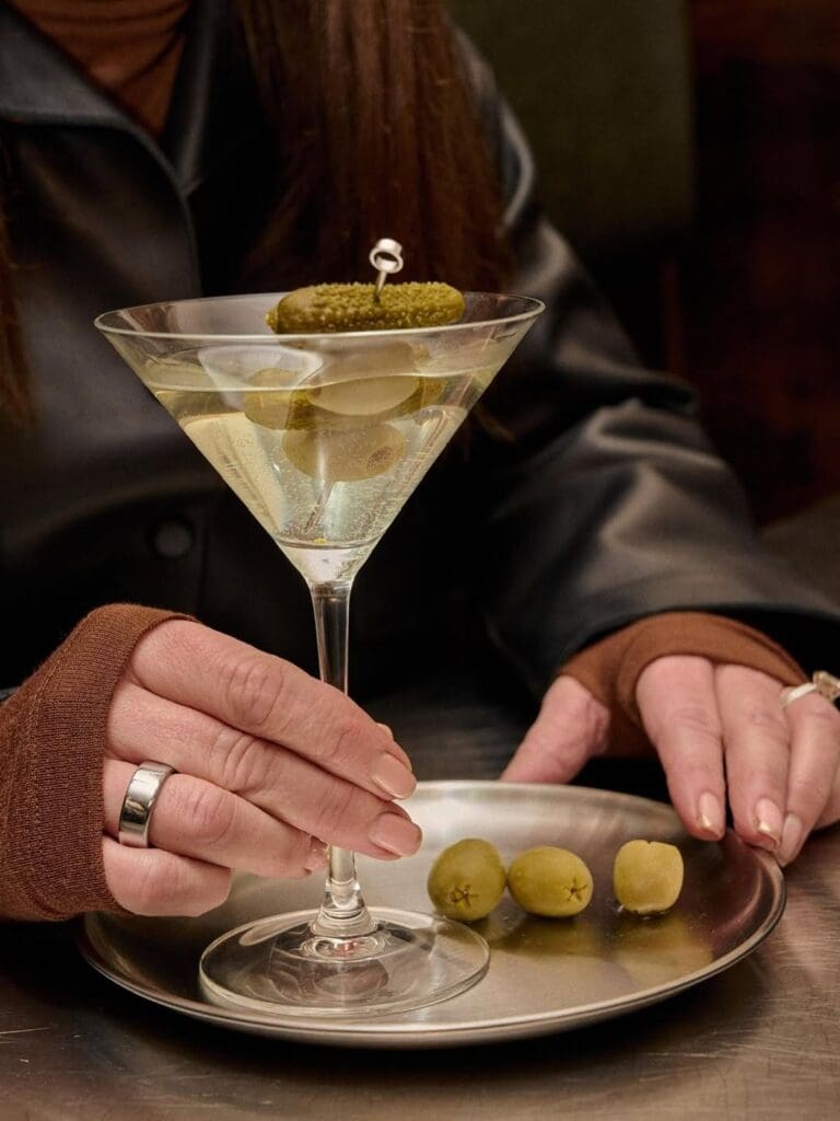 A close-up of a dirty martini garnished with a cornichon on a cocktail pick, served in a classic martini glass on a silver plate with three olives on the side. A woman in a leather jacket holds the glass at The Uncommon in Newcastle.