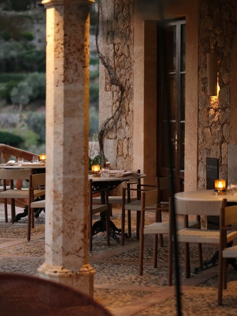 The candlelit outdoor terrace at De Tokio a Lima in Palma at dusk, with intimate tables set between stone columns and rustic stone walls, woven-seat wooden chairs, climbing vines, warm wall lighting and green hillside views in the background.