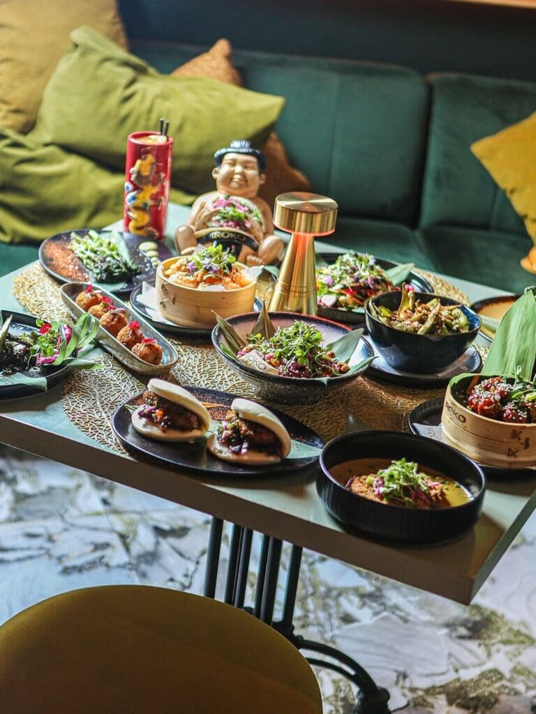 A table full of Asian-inspired sharing dishes at Iroko in Palma, including bao buns, crispy fried bites, bamboo steamer baskets, noodle bowls and vibrant salads topped with microherbs and edible flowers. The table is set against a teal velvet sofa with green and mustard cushions, with a gold table lamp, a decorative sumo wrestler figurine and a dragon-print cocktail glass adding to the maximalist feel.
