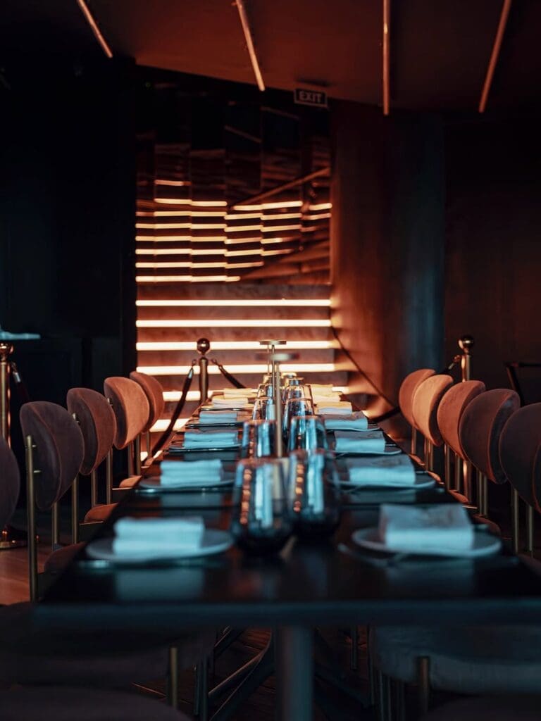 A long dark dining table set for a group at Lío in Palma, with smoked glass wine glasses, folded napkins and place settings lit by warm amber strip lighting on the back wall, giving the room a sleek, nightclub-meets-restaurant feel.