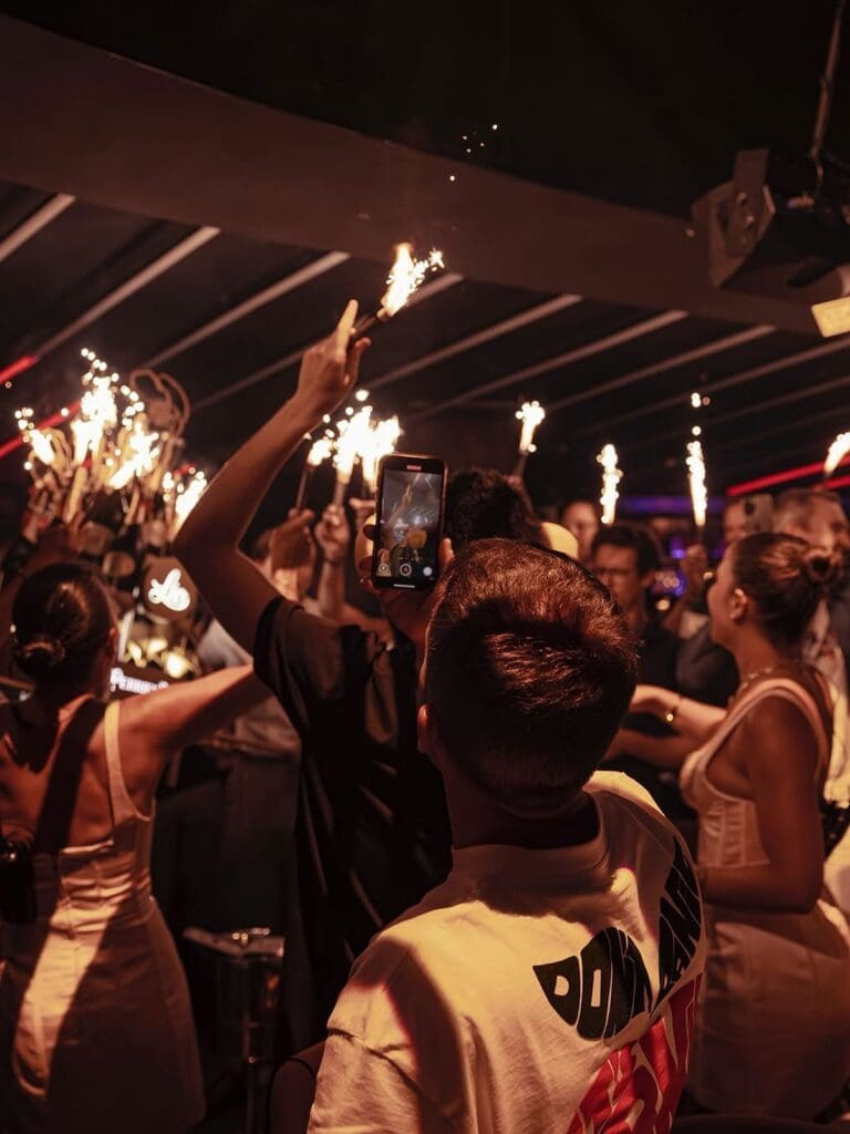 A crowd at Lío in Palma holding up sparklers and filming on their phones during a late-night party moment, with warm orange light filling the room and people dancing in the background