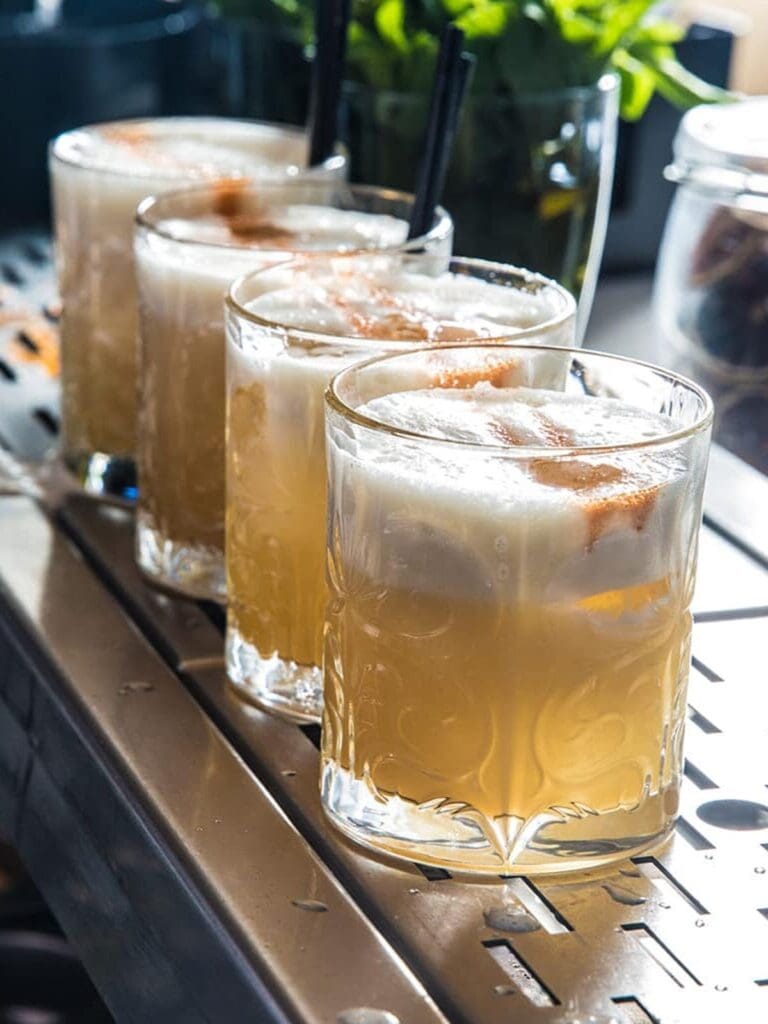 A row of four golden cocktails with frothy white tops and black straws lined up on the bar at Siso Beach in Palma, served in embossed glass tumblers with fresh herbs visible in the background.