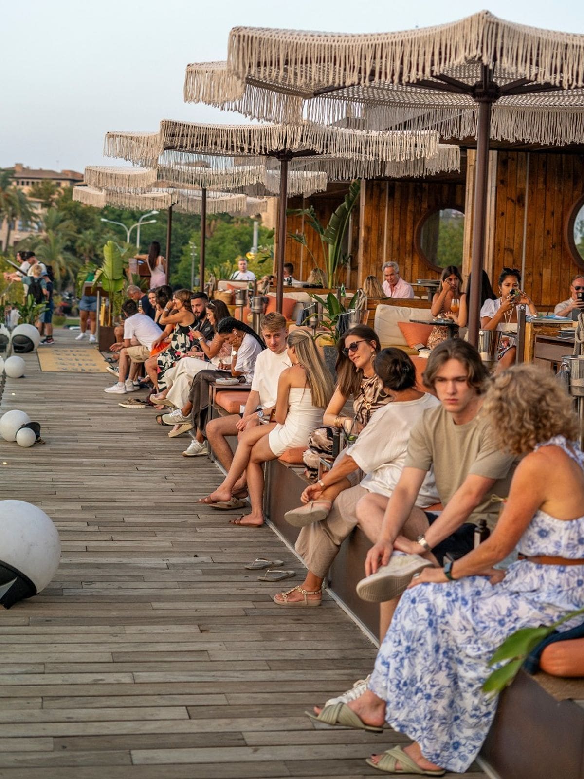 Groups of people sitting along the wooden boardwalk terrace at Sky Nudos in Palma during golden hour, with fringed parasols overhead, a timber-clad bar in the background, and a relaxed, buzzy holiday atmosphere.