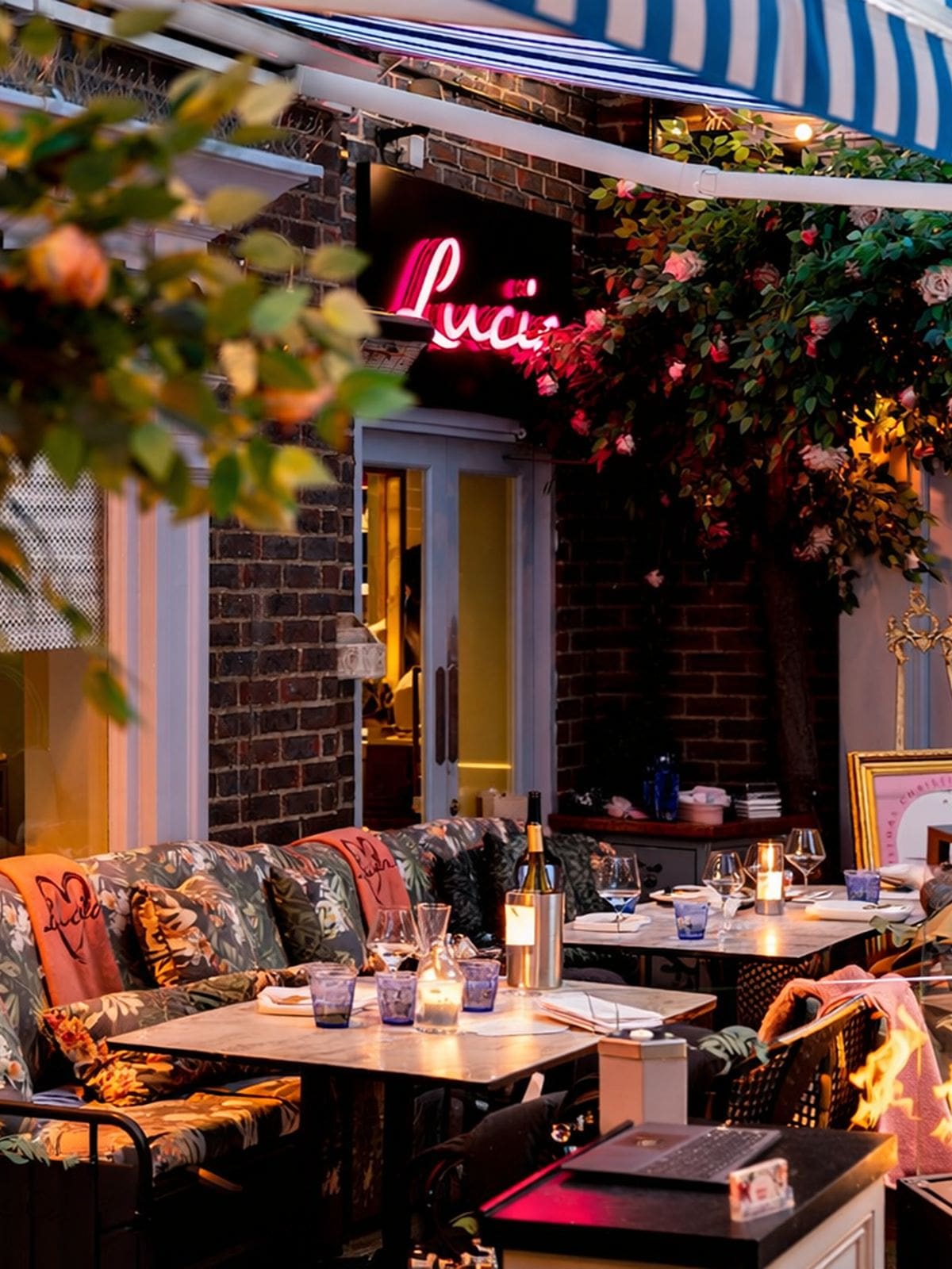 The outdoor terrace at Lucia's in York at dusk, with a pink neon Lucia sign glowing above the entrance, floral print cushioned seating, candlelit tables set with wine glasses and a bottle of wine, a blue and white striped awning overhead, and climbing roses on the exposed brick wall.