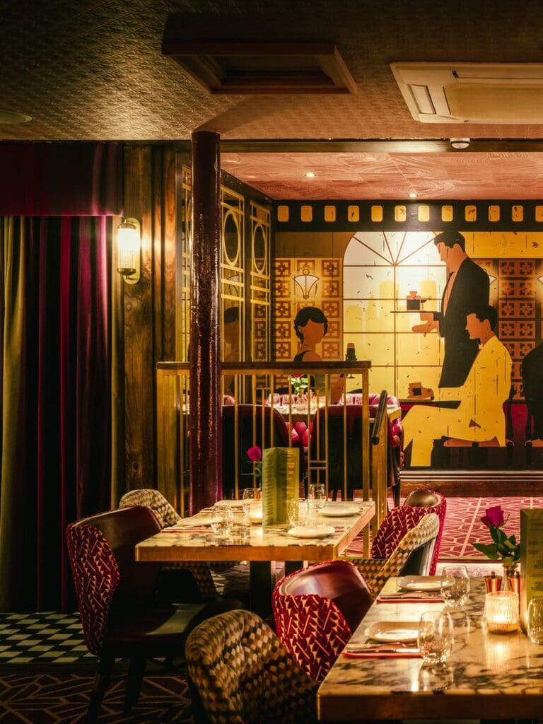 The moody, art deco-inspired dining room at Nola in York, with marble-topped tables set with wine glasses and single stem roses, patterned upholstered chairs in deep reds and golds, velvet curtains, art deco wall sconces, and a large vintage-style mural of a waiter serving cocktails on the back wall.