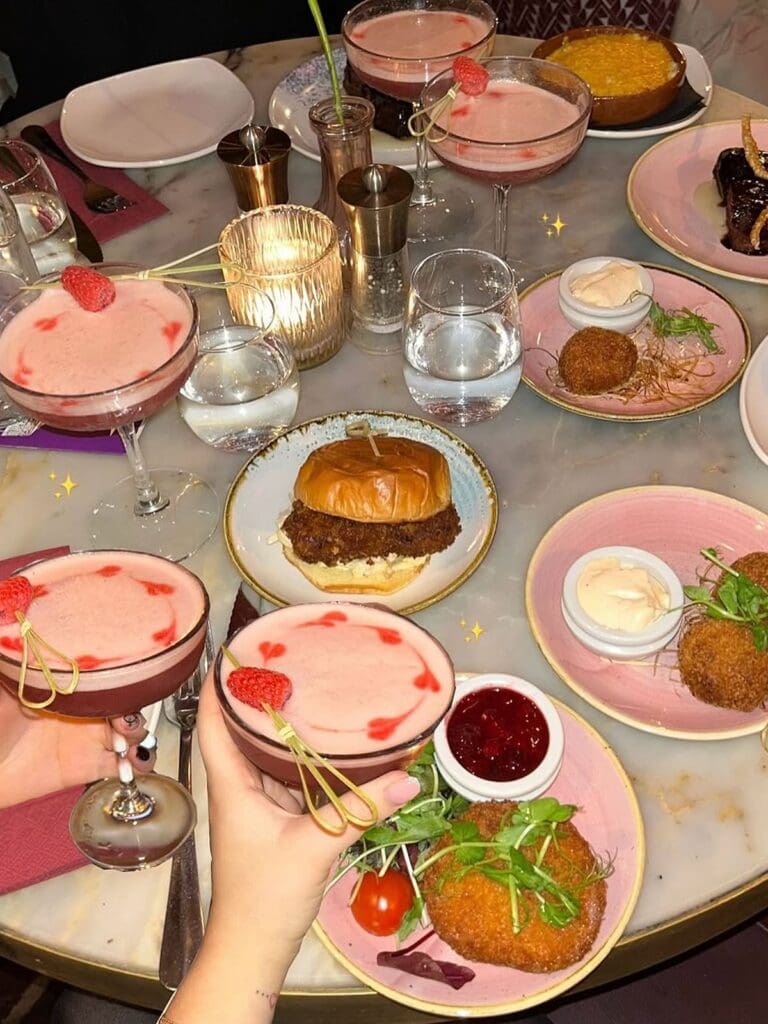 An overhead shot of a marble table at Nola in York covered in pink raspberry cocktails garnished with hearts and fresh raspberries, a brioche burger, golden fried croquettes on pink plates with dipping sauces, a crème brûlée and a candle glowing in the middle. A hand reaches in to pick up one of the cocktails.