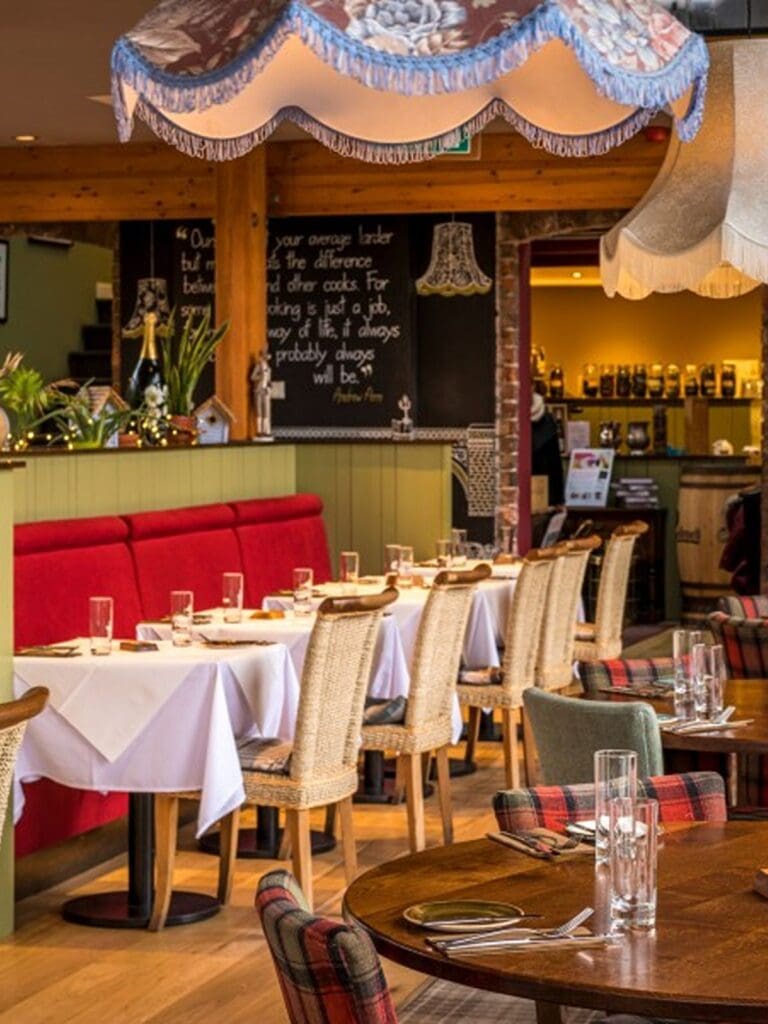 The dining room at Star Inn The City in York, with white tablecloth-covered tables, red banquette seating, wicker-backed chairs, tartan-upholstered seats, vintage fringed lampshades overhead, a chalkboard quote on the wall, green tongue-and-groove panelling, and wooden beams giving the space a cosy, country pub feel.