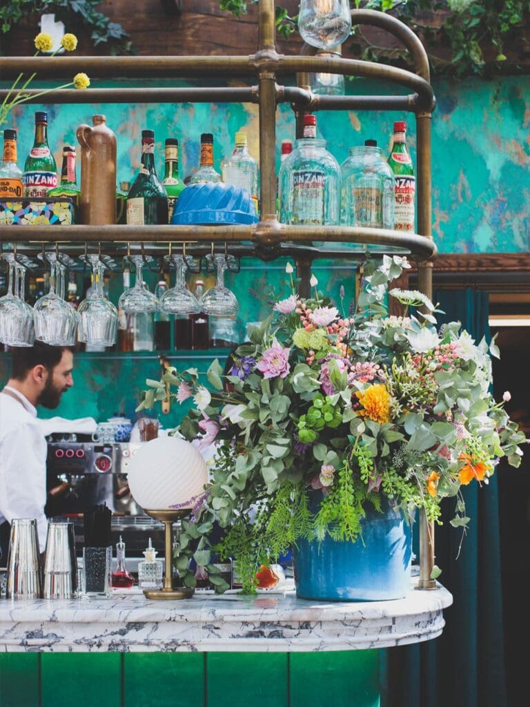Paris bar interior with floral arrangements, hanging glassware and a marble counter, creating a lush, boozy-lunch setting for a girls’ weekend.