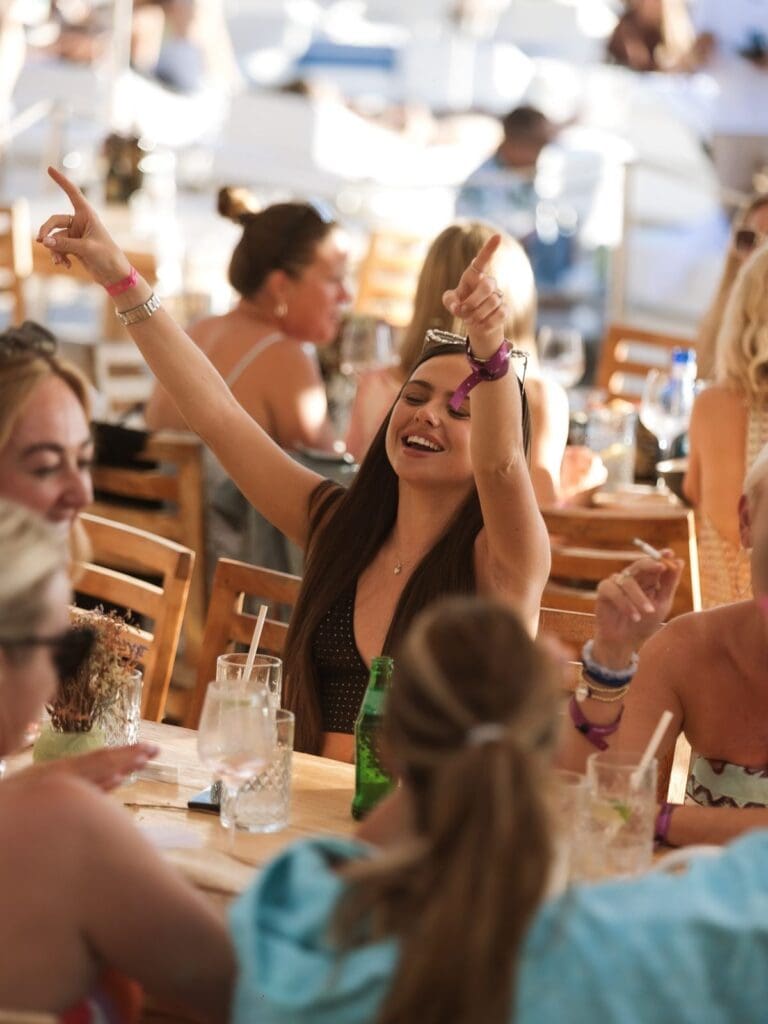 Girls’ hen party at Blue Marlin Ibiza raising their arms and dancing at a beachfront dinner table in golden evening light.