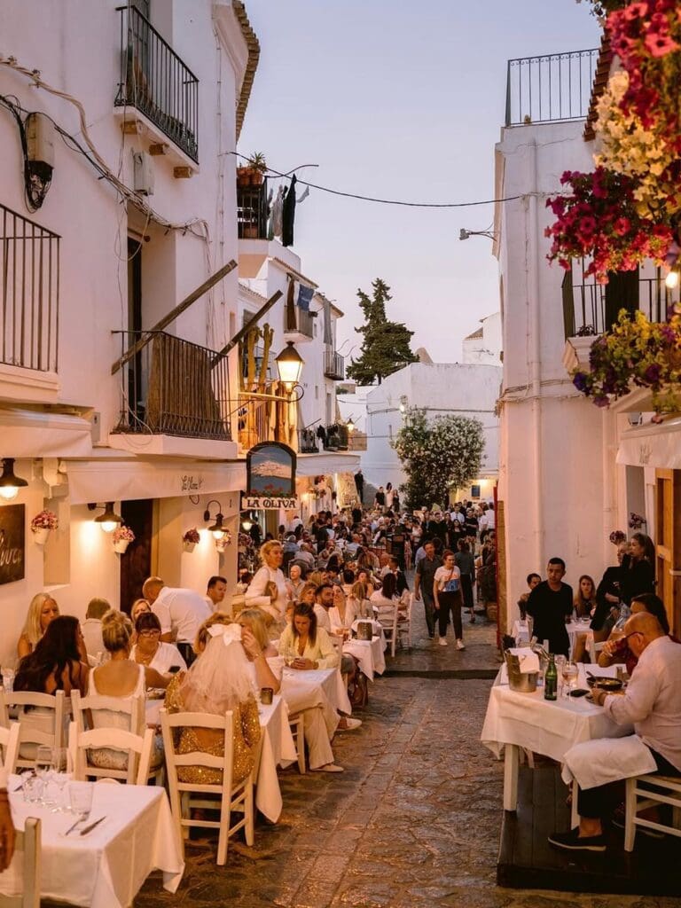 Outdoor evening dining at La Oliva in Ibiza, with white-clothed tables lining a narrow, whitewashed street filled with people enjoying dinner in the warm glow of lanterns. The lively, buzzy atmosphere and a bride-to-be in a veil make it perfect for hen weekends or stylish girls weekends in Ibiza.
