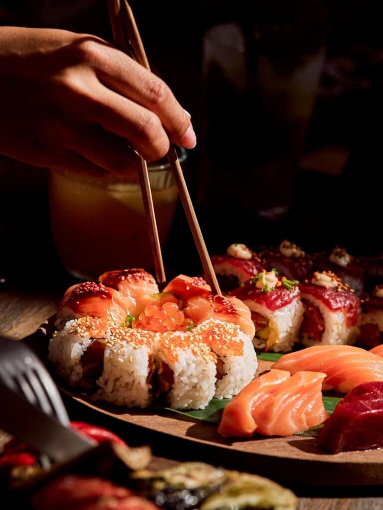 Sushi platter and sashimi at Playa Soleil Ibiza during a hen weekend dinner, with chopsticks reaching in under warm evening lighting.