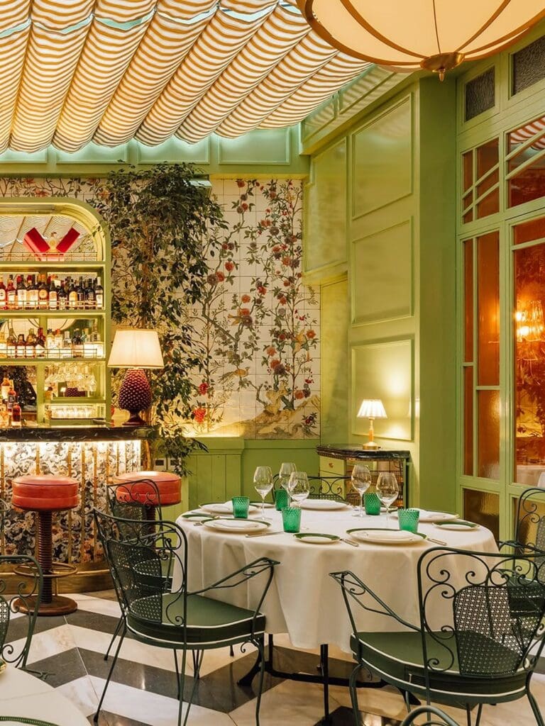 Elegant green dining room at Rocco Lisbon featuring hand-painted botanical tiles, striped ceiling canopy, wrought-iron chairs and a round table set with white linen and green glassware for a hen weekend dinner.