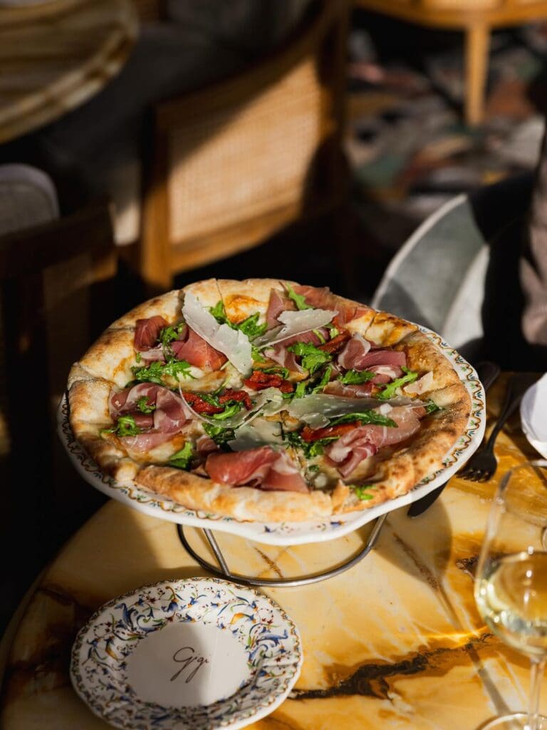A wood-fired pizza topped with prosciutto, rocket, sun-dried tomatoes and parmesan shavings, served on a raised stand on a hand-painted Italian-style plate at Gigi's in Paris, with a branded Gigi side plate, a glass of white wine and cane-backed chairs visible in the background.